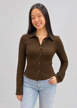 A woman wearing a brown ribbed collared button-up top with slight bell sleeves, paired with blue denim jeans.